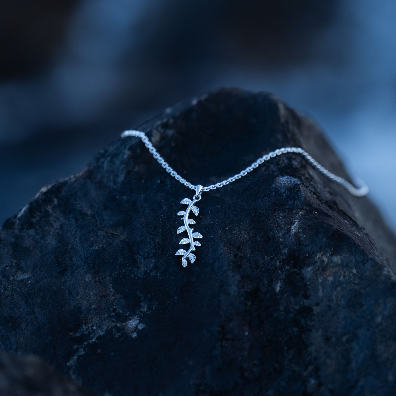 A silver leaf pendant necklace with delicate botanical details laying on dark rock