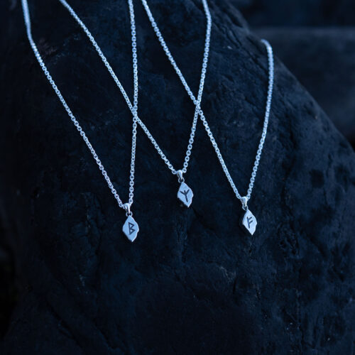 Three silver rune necklaces with different runes laying on a dark rock